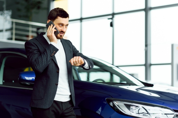 man looking at watch while on the phone in front of his blue car depicting factors in choosing auto glass repair shop